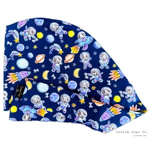 Könnte beinhalten: Eine blaue Stoff-Operationsmütze mit einem sich wiederholenden Muster aus Cartoon-Hunden in Astronautenanzügen, Planeten, Sternen und Raketen. Der Text "Custom Caps Co." ist in der rechten unteren Ecke der Mütze aufgedruckt.