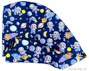 Könnte beinhalten: Eine blaue Stoff-Operationsmütze mit einem sich wiederholenden Muster aus Cartoon-Hunden in Astronautenanzügen, Planeten, Sternen und Raketen. Der Text "Custom Caps Co." ist in der rechten unteren Ecke der Mütze aufgedruckt.