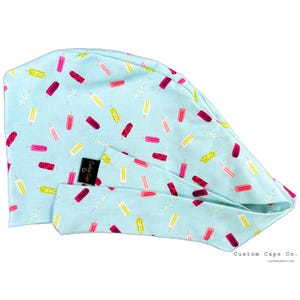 May include: A light blue surgical scrub cap with a pattern of colorful popsicle illustrations. The popsicles are in shades of pink, red, and yellow. The cap has a black label that says "Custom Caps Co."