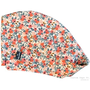 May include: A floral scrub cap with a cream background and a repeating pattern of pink roses, blue-green leaves, and yellow-brown accents. The brand name "Custom Caps Co." is visible on a small black tag. The cap is designed for medical professionals.