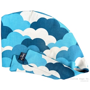 May include: A blue and white patterned surgical cap with a tie closure. The pattern features a repeating design of clouds. The cap has a label that reads "Custom Caps Co."
