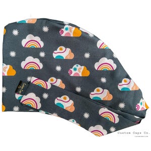 May include: A dark grey scrub cap featuring a pattern of white clouds, rainbows, and starbursts. The clouds have orange, pink, and teal accents. The cap has a matching tie and the text "Custom Caps Co."