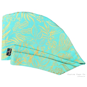 May include: A turquoise blue fabric scrub cap with a gold leaf pattern. The cap has a small black label with the text "Custom Caps Co."
