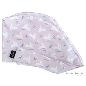 Peut inclure: Une charlotte chirurgicale rose avec un motif de lapin blanc. La charlotte a une attache à l'arrière et le texte "Custom Caps Co. customcapsco.com" est imprimé en bas à droite.