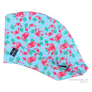 May include: A light blue surgical cap with a pattern of red crabs on a light blue background. The cap has a tie at the back. The text "Custom Caps Co." is printed on the bottom right corner of the image.