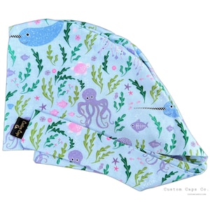 Peut inclure: Une toque chirurgicale bleu clair et un bandeau assorti, ornés de créatures marines telles que des narvals, des pieuvres et des poissons. Le motif comprend des algues vertes, des étoiles de mer roses et d'autres éléments aquatiques. La toque et le bandeau sont en tissu doux.