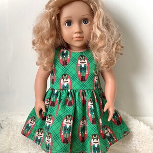May include: A doll wearing a green dress with a repeating nutcracker soldier pattern. The dress has a plaid design and red trim. The doll has blonde curly hair and is standing on a white surface.