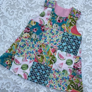 May include: A patchwork dress featuring a variety of floral, watermelon, and patterned fabrics. The sleeveless dress has a pink lining and is adorned with small purple buttons on the shoulders. The dress is designed for a child.