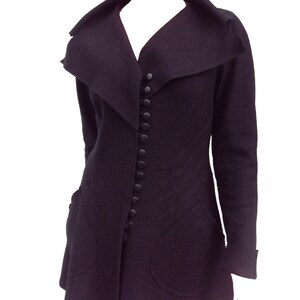 May include: A black long-sleeved jacket with a large, asymmetrical collar and a row of buttons down the front. The jacket has two large pockets on the front and a fitted silhouette.