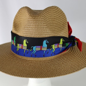 May include: A brown straw hat with a black and blue ribbon featuring a colorful horse design. The ribbon is tied in a bow at the top of the hat.