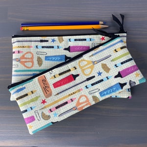 May include: Two rectangular fabric pencil cases with black zippers, featuring a cream-colored grid pattern printed with colorful illustrations of scissors, markers, pencils, erasers, and stars. Two pencils rest on top of the larger case.