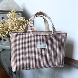 May include: A quilted tote bag featuring a brown and white gingham check pattern. The bag has two matching fabric handles and a small white rectangular label on the front with the text Beyond Sassy Handmade printed in black lettering.