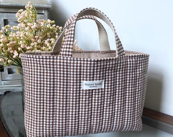 May include: A quilted tote bag featuring a brown and white gingham check pattern. The bag has two fabric handles and a white rectangular label on the front with the text Beyond Sassy Handmade printed in black. The interior lining is beige.