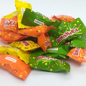 Könnte beinhalten: Ein Haufen einzeln verpackter Bonbons in grünen, gelben und orangen Verpackungen. Die Bonbons sind mit "ШИПУЧКА" und "ROSHEN" gekennzeichnet. Die Verpackungen haben ein Muster aus roten und gelben Punkten und einem Sternenausbruch.