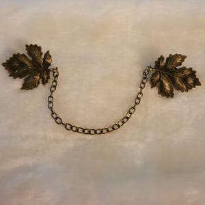 May include: A vintage-style bronze-toned leaf brooch with a chain. The brooch features two detailed leaf-shaped ornaments connected by a delicate chain. The leaves have intricate veining and a warm, antique finish, suggesting a classic or nature-inspired design.