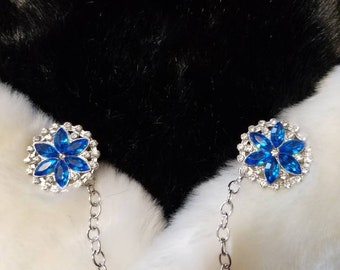 May include: A pair of blue rhinestone flower brooches connected by a silver chain. The brooches are on a white and black fur background.