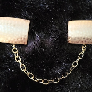 May include: A pair of gold-toned metal collar clips connected by a gold chain. The clips have a textured, hammered finish.