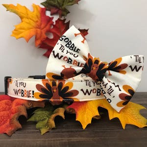 Może przedstawiać: Biały obroża dla psa z nadrukiem brązowego i pomarańczowego indyka i tekstem "Gobble 'Til You Wobble!" Pokazano r&oacute;wnież pasującą muszkę z tym samym nadrukiem i tekstem.