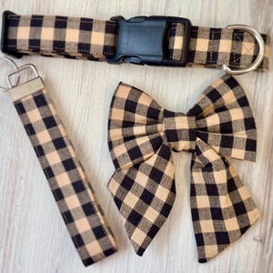May include: A black and tan gingham dog collar, leash, and bow tie. The collar has a black plastic buckle. The leash is attached to a silver key ring. The bow tie is tied in a classic bow shape.