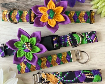 May include: Three different dog collars with Mardi Gras themed fabric. The collars are purple, green, and gold with glitter and beads. The collars are paired with matching flower brooches.
