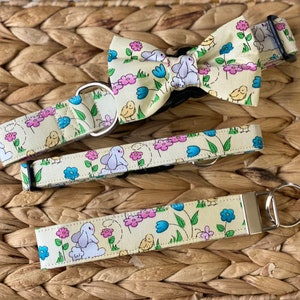 May include: A set of three matching pet accessories featuring a light yellow background with a repeating pattern of white bunnies, pink and blue flowers, and yellow chicks. The set includes a collar, a bow tie, and a key chain.