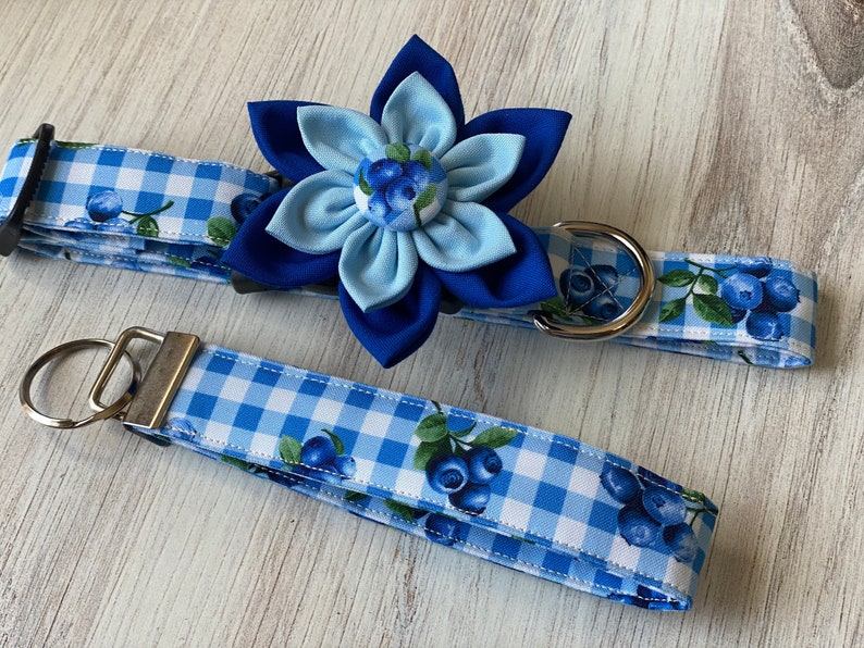 May include: Blue and white gingham dog collar with a blue flower and blueberry print. The collar is attached to a matching blue and white gingham key chain.