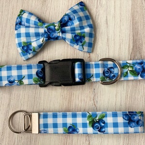 Op de afbeelding: Een blauwe en witte gingham hondenriem met een zwarte gesp en een bijpassende strik met bosbessenprint. De riem is 2,5 cm breed en de strik is 7,5 cm breed.