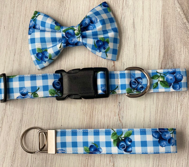 Op de afbeelding: Een blauwe en witte gingham hondenriem met een zwarte gesp en een bijpassende strik met bosbessenprint. De riem is 2,5 cm breed en de strik is 7,5 cm breed.