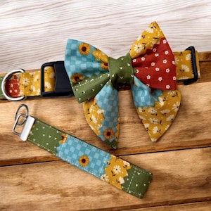 May include: A pet collar featuring a large fabric bow tie and a matching keychain strap. The fabric displays a patchwork pattern of sunflowers, white daisies, and blue checkered squares in shades of yellow, green, red, and light blue with a black buckle.