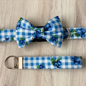 Op de afbeelding: Blauw-witte gingham hondenhalsband en bijpassende strik met een blauwe bessenprint. De halsband heeft een zilveren metalen D-ring en de strik heeft een zwarte verstelbare band. De set bevat ook een bijpassende riem met een zilveren sleutelhanger.