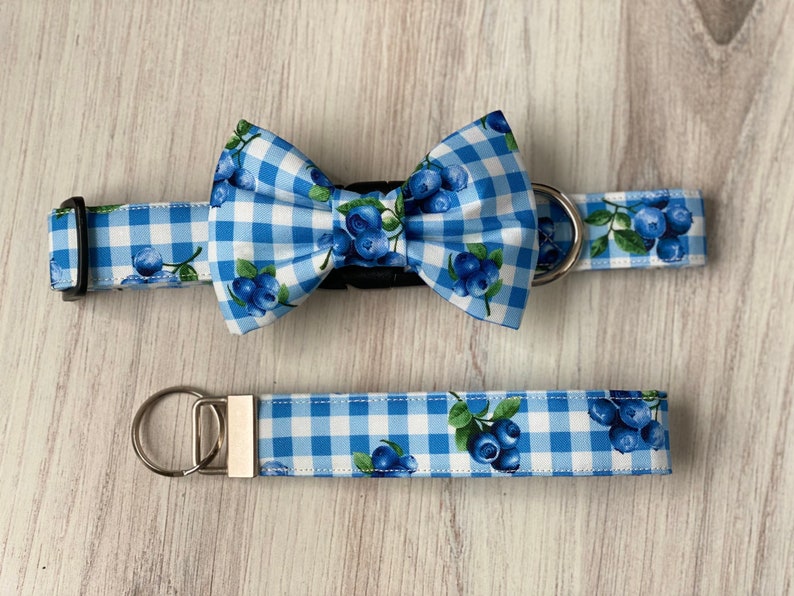 Op de afbeelding: Blauw-witte gingham hondenhalsband en bijpassende strik met een blauwe bessenprint. De halsband heeft een zilveren metalen D-ring en de strik heeft een zwarte verstelbare band. De set bevat ook een bijpassende riem met een zilveren sleutelhanger.