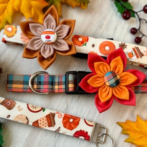 May include: Three dog collars with different patterns and fabric. One collar is plaid, one is white with a donut and apple pattern, and one is white with a fall-themed pattern. Each collar has a fabric flower attached. The flowers are in shades of orange, red, and brown.