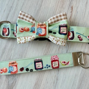 May include: A green and white dog collar with a matching bow tie. The collar and bow tie feature a pattern of jars of jam, cherries, and a spoon. The bow tie has a gingham pattern on the back. The collar has a metal buckle and a matching lead.