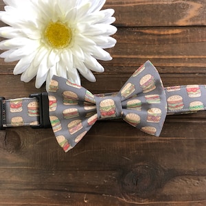 May include: A grey bow tie with a pattern of colourful hamburgers. The bow tie is made of fabric and has a black adjustable strap.