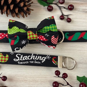 May include: A black dog collar with a red and white polka dot pattern and a matching bow tie with a green and red elf hat and mustache pattern. The collar has a silver buckle and a silver D-ring. The bow tie has a black background with a white snowflake and the text "Stacking Through The Snow..." on the back.
