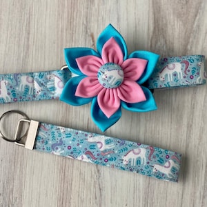 May include: A blue and pink fabric flower with a unicorn button in the center. The flower is attached to a blue fabric collar with a white unicorn print. A matching blue fabric key chain with a white unicorn print is also included.