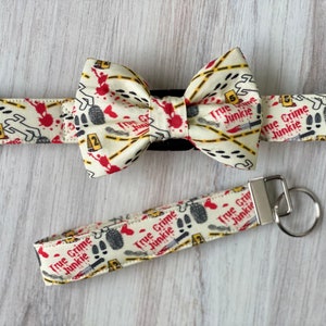 May include: A white bow tie and matching dog collar with a "True Crime Junkie" print. The print features red blood splatter, crime scene tape, and footprints. The bow tie and collar are attached to a silver key chain.