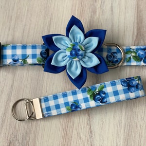 May include: Blue and white gingham dog collar with a blue flower charm and matching lead. The flower charm has a blue centre with white and blue blueberry accents.