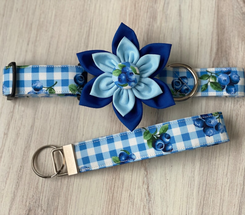 May include: Blue and white gingham dog collar with a blue flower charm and matching lead. The flower charm has a blue centre with white and blue blueberry accents.