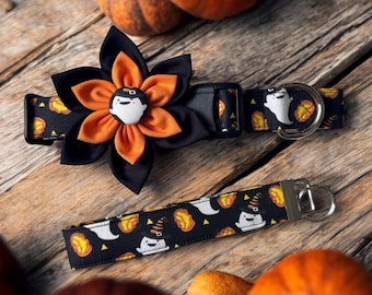 May include: A black pet collar featuring a pattern of white ghosts and orange pumpkins, adorned with a large layered fabric flower in black and orange with a central ghost button. A matching fabric keychain strap with the same ghost pattern.