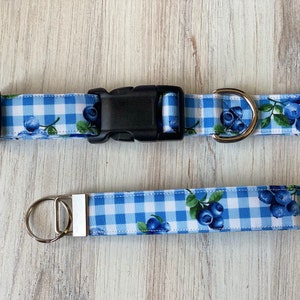 Op de afbeelding: Blauw en wit gingham hondenhalsband en bijpassende riem met een blauwe bessenprint. De halsband heeft een zilveren gesp en een zilveren D-ring. De riem heeft een zilveren splitring.