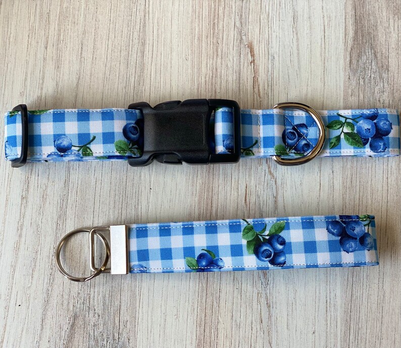 Op de afbeelding: Blauw en wit gingham hondenhalsband en bijpassende riem met een blauwe bessenprint. De halsband heeft een zilveren gesp en een zilveren D-ring. De riem heeft een zilveren splitring.