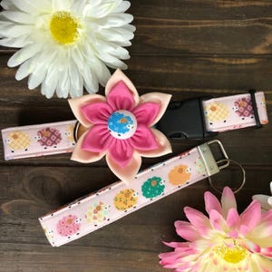 May include: A pink and white flower with a blue and white button center is attached to a pink and white fabric dog collar with a black buckle. A matching pink and white fabric key chain with a silver key ring is also shown.