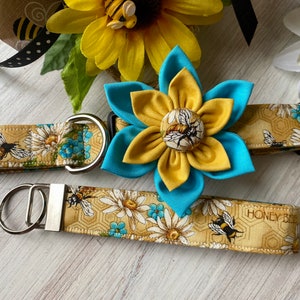 May include: A yellow and blue fabric flower with a bee design in the centre. The flower is attached to a brown and yellow fabric dog collar with a matching lead. The lead has a silver key ring and the text "Honey Bee" printed on it.