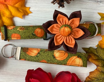 May include: A green and brown patterned dog collar with a matching leash and a brown and orange fabric flower accent. The collar and leash feature a pumpkin print.