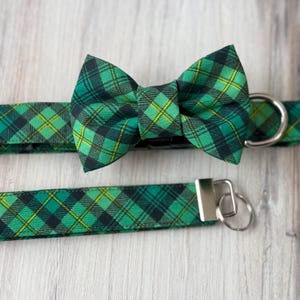May include: A green and black plaid dog collar with a matching bow tie. The collar has a silver buckle and a silver D-ring. The bow tie is tied in a classic bow shape.