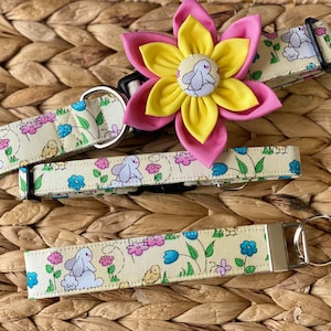 May include: A dog collar, a matching key fob, and a flower accessory. The collar and key fob feature a light yellow fabric with a repeating pattern of bunnies, flowers, and chicks. The flower accessory has pink and yellow petals with a bunny center.