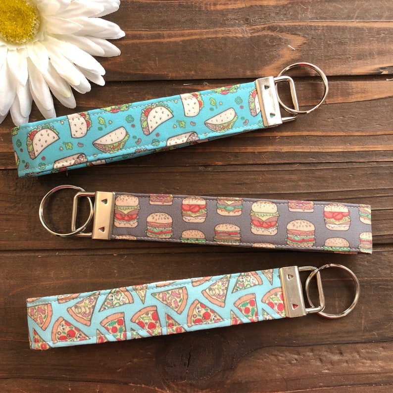 Op de afbeelding: Drie sleutelhangers met verschillende voedselontwerpen. De eerste sleutelhanger is blauw met een witte achtergrond en heeft taco's. De tweede sleutelhanger is grijs met een witte achtergrond en heeft hamburgers. De derde sleutelhanger is blauw met een witte achtergrond en heeft pizzapunten.