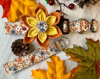 Pode incluir: Um conjunto de acessórios para cães em cores outonais. Uma flor de tecido marrom e amarelo com um botão cinza no centro está presa a uma coleira de cão bege com estampa floral. Também está incluída uma guia com estampa floral combinando com um chaveiro prateado.