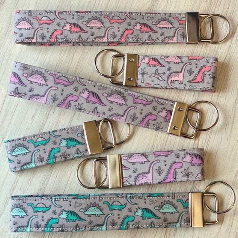 May include: Five keychains with different dinosaur patterns. The keychains are made of fabric with a gray background and feature pink, green, and blue dinosaurs. The keychains have gold metal rings and clasps.